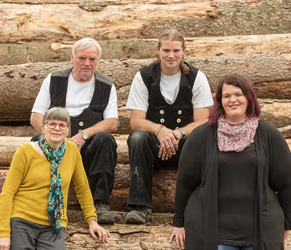 Vier Personen stehen vor gestapelten Baumstämmen.